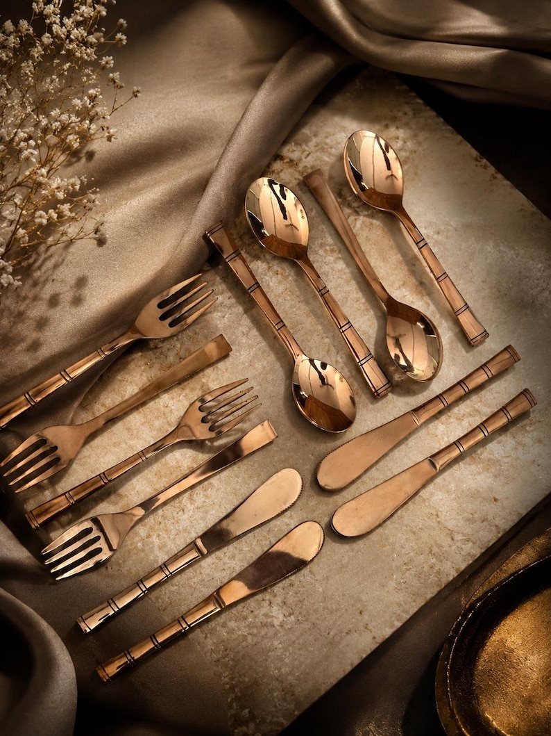 May include: A set of rose gold-toned flatware including three forks, two spoons, and three knives. Each piece features a polished metallic finish and a distinct handle design with three horizontal etched lines near the neck of the utensil.