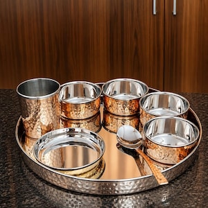 Puede incluir: Una bandeja de servicio redonda que contiene un vaso alto, cuatro cuencos pequeños, un plato poco profundo y una cuchara. Cada pieza presenta un acabado exterior de cobre martillado y una superficie interior plateada y lisa.