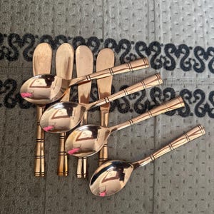 May include: A set of four copper-colored metal spoons and four matching flat-edged knives arranged in a row. Each piece features a polished finish and a handle decorated with three distinct horizontal bands near the end of the stem.