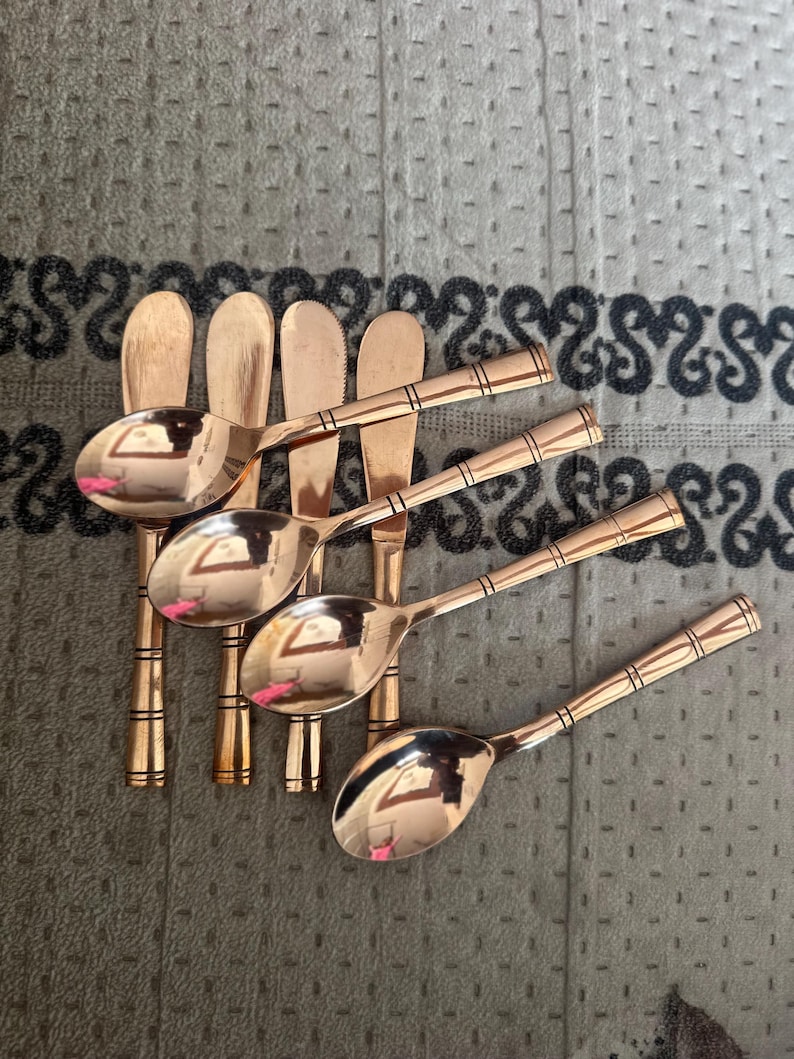 May include: A set of four copper-colored metal spoons and four matching flat-edged knives arranged in a row. Each piece features a polished finish and a handle decorated with three distinct horizontal bands near the end of the stem.