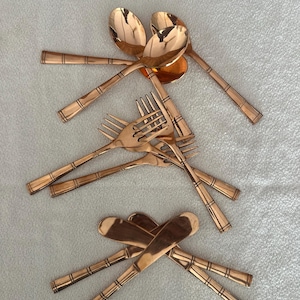 May include: A collection of copper-colored flatware including spoons, forks, and knives. Each piece features a polished metallic finish and a handle decorated with three distinct horizontal bands. The utensils are arranged in three separate groups.