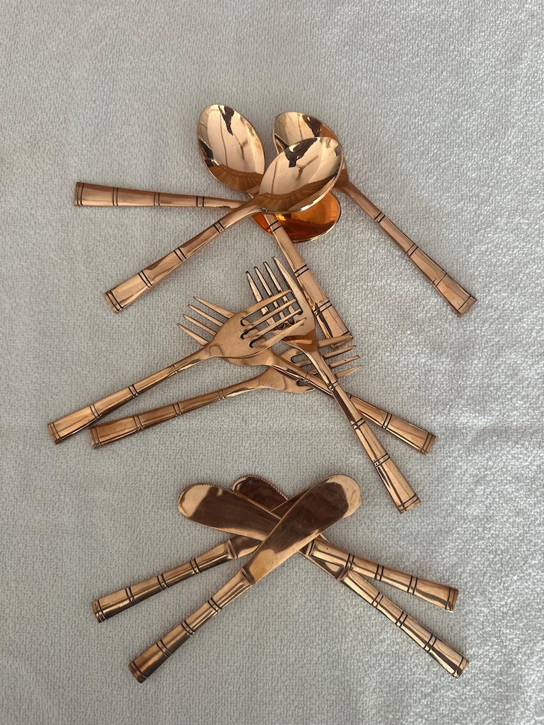 May include: A collection of copper-colored flatware including spoons, forks, and knives. Each piece features a polished metallic finish and a handle decorated with three distinct horizontal bands. The utensils are arranged in three separate groups.
