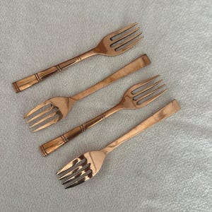 May include: A set of four copper-colored metal forks arranged diagonally. Two forks feature a three-tine design with a decorative banded handle, while the other two forks have a smooth, flat handle design and four tines, all with a metallic finish.