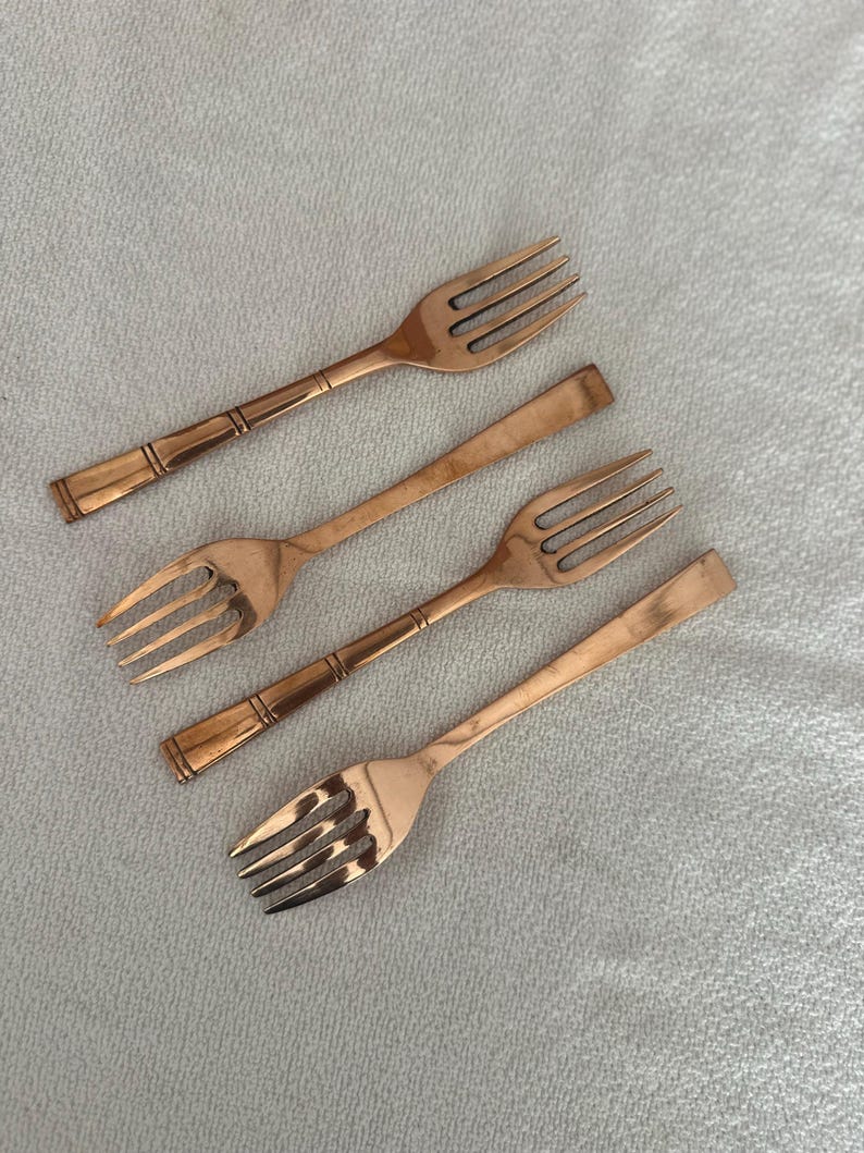 May include: A set of four copper-colored metal forks arranged diagonally. Two forks feature a three-tine design with a decorative banded handle, while the other two forks have a smooth, flat handle design and four tines, all with a metallic finish.