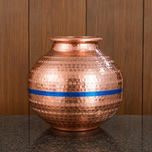 Puede incluir: Una olla de cobre martillado de forma redonda con una banda horizontal ancha de color azul vibrante que rodea su centro. El recipiente tiene un cuello corto y ensanchado y un borde plano y liso, con una superficie texturizada por pequeñas hendiduras.