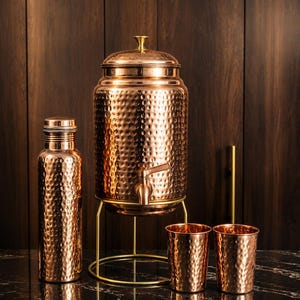 May include: A hammered copper water set consisting of a tall cylindrical water dispenser with a spigot on a gold metal stand, a matching water bottle with a screw-top lid, and two matching hammered copper tumblers with a polished finish.