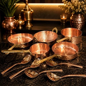 May include: A collection of hammered copper cookware including three round pans with brass handles, a deep bowl with two brass handles, and four matching hammered copper spoons. The cookware features a polished finish and a distinct textured surface.