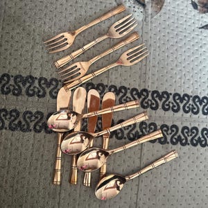 May include: A collection of rose gold-toned metal cutlery consisting of four forks and five spoons. Each piece features a polished finish and a handle designed with three distinct horizontal bands. The utensils are arranged in a cluster.