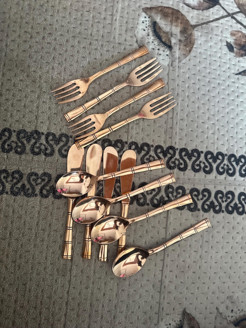 May include: A collection of rose gold-toned metal cutlery consisting of four forks and five spoons. Each piece features a polished finish and a handle designed with three distinct horizontal bands. The utensils are arranged in a cluster.
