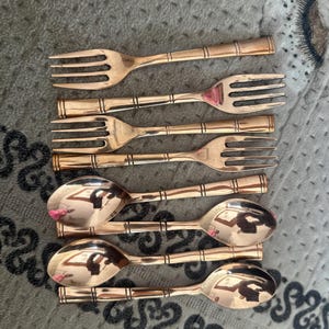 May include: A set of four copper-toned metal forks and four matching spoons arranged in two rows. Each piece of flatware features a smooth, polished finish and a distinct handle design with three horizontal bands etched into the metal surface.