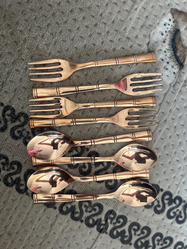 May include: A set of four copper-toned metal forks and four matching spoons arranged in two rows. Each piece of flatware features a smooth, polished finish and a distinct handle design with three horizontal bands etched into the metal surface.
