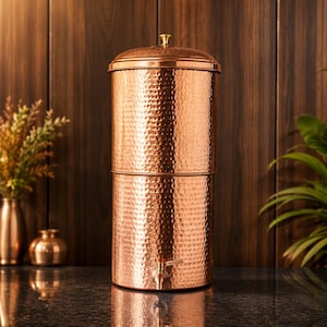 May include: A cylindrical copper water dispenser featuring a hammered texture throughout its exterior surface. The vessel includes a removable lid with a small gold-toned knob at the top and a functional metal spigot positioned near the base.