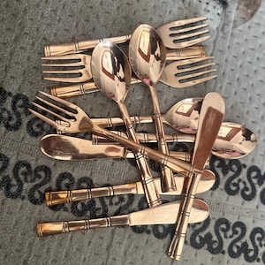 May include: A collection of copper-toned metal flatware featuring a bamboo-inspired design on the handles. The set includes several forks, spoons, and knives with a polished, reflective finish, all arranged in a layered, overlapping pile.