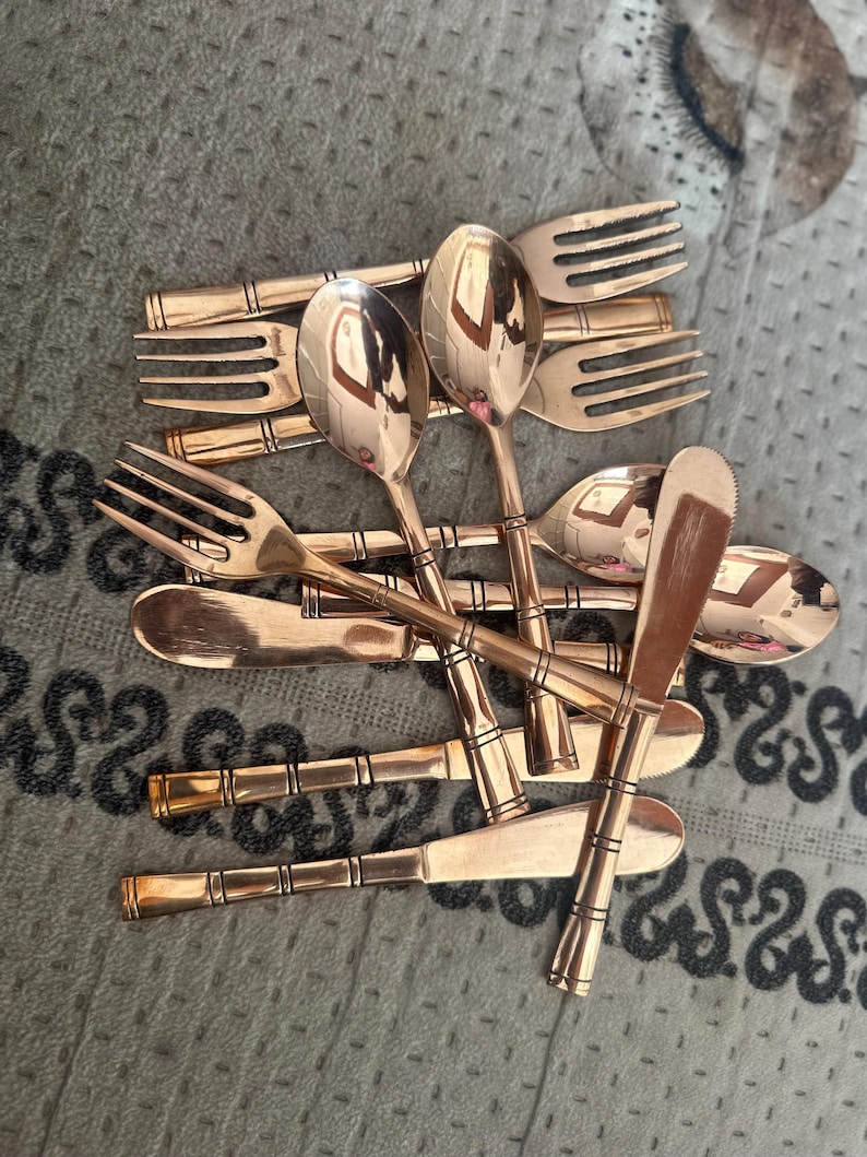 May include: A collection of copper-toned metal flatware featuring a bamboo-inspired design on the handles. The set includes several forks, spoons, and knives with a polished, reflective finish, all arranged in a layered, overlapping pile.