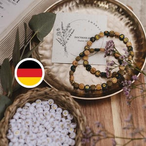 Könnte beinhalten: Zwei kreisförmige Armbänder aus kleinen, runden Perlen mit mattem Finish in Braun-, Schwarz- und Beigetönen. Jede Perle ist mit einem goldenen Buchstaben versehen, die zusammen Wörter bilden. Die Armbänder liegen auf einem goldenen Metalltablett.
