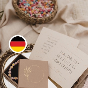 Könnte beinhalten: Eine quadratische braune Pappschachtel mit einem goldenen Blumen-Logo, die ein rosa Perlenarmband mit gelben Buchstabenperlen enthält. Daneben liegt eine cremefarbene Karte mit Text auf einem ovalen Spiegeltablett mit goldenem Rand.