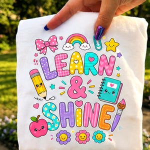 Puede incluir: Una camiseta blanca con un gráfico colorido que incluye el texto "LEARN & SHINE". El diseño presenta un lápiz amarillo, un cuaderno azul verdoso, una manzana roja, un lazo rosa, un arcoíris, una estrella y un pincel, todos con caras sonrientes.