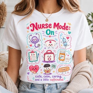 May include: A white t-shirt featuring a colorful graphic with the text "Nurse Mode: On" and "cute, calm, caring, and a little caffeinated." The design includes cartoon illustrations of a stethoscope, nurse cap, syringe, clipboard, medical kit, pill bottle, heart, coffee cup, and bandage.