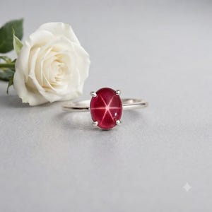 May include: A silver ring featuring a central oval-shaped cabochon-cut red star ruby gemstone with a distinct six-rayed star pattern. The gemstone is held in a four-prong setting on a polished silver band, positioned next to a white rose.