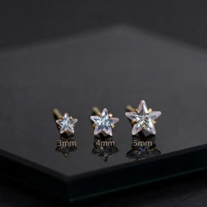 May include: Three star-shaped stud earrings with clear, faceted stones set in gold-toned metal. The earrings are arranged by size, labeled as 3mm, 4mm, and 5mm, each featuring a pointed star design with a central stone and metallic prongs.