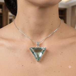 May include: A silver necklace featuring a large, triangular, faceted light blue gemstone pendant. The pendant is held by a decorative silver bail and hangs from a delicate silver box chain that rests against the collarbone area.