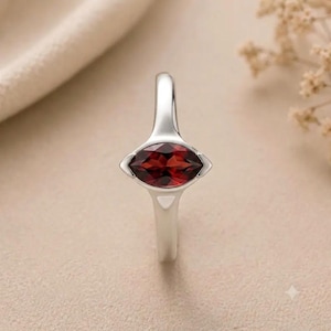 May include: A silver ring featuring a marquise-cut deep red garnet gemstone set horizontally in a smooth metal band. The setting has a pointed, geometric design that tapers into the band, showcasing the dark crimson facets of the stone.