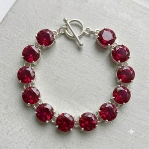 May include: A silver-toned bracelet featuring a series of round, faceted, deep red stones set in individual metal frames. The bracelet is secured with a circular toggle clasp mechanism at the top, connecting the two ends of the circular chain.