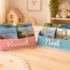 Könnte beinhalten: Zwei Schreibtisch-Bücherregale aus Kunststoff, eines in Rosa mit dem Namen Hannah und eines in Hellblau mit dem Namen Noah, die jeweils mehrere Kinderbücher enthalten, darunter Titel wie Conni geht zum Zahnarzt, Conni macht einen Ausflug, Conni entdeckt die Bücher und Conni geht einkaufen.