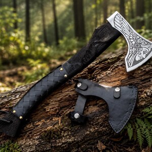 Può includere: Un'accetta con manico in legno scuro e testurizzato, avvolto in pelle intrecciata nera e fissato con rivetti in ottone. La testa dell'ascia in metallo argentato presenta un'intricata incisione di una figura alata con fodero nero.