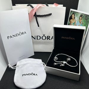 Könnte beinhalten: Ein silbernes Pandora-Schlangenkettenarmband mit herzförmigen Charms in einer weißen Schmuckschatulle, begleitet von einer weißen Geschenktüte, einer weißen Papiertasche und einem weißen Stoffbeutel, alle mit dem schwarzen Pandora-Logo.