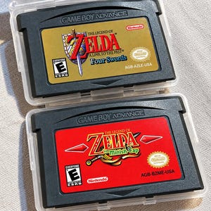 May include: Two Game Boy Advance video game cartridges in clear plastic cases. The top cartridge has a gold label for The Legend of Zelda: A Link to the Past & Four Swords. The bottom cartridge has a red label for The Legend of Zelda: The Minish Cap.