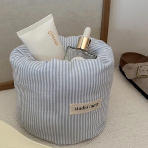Op de afbeelding: Een cilindervormig opbergzakje van stof met een verticaal gestreept patroon in lichtblauw en wit. Het zakje heeft een klein rechthoekig label aan de voorzijde met de tekst studio mosy in het zwart gedrukt. Het bevat een witte tube en een glazen pipetflesje.