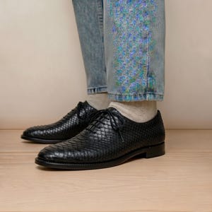 Puede incluir: Un par de zapatos oxford negros con cordones y un patrón texturizado similar a la piel de serpiente. Los zapatos tienen un tacón bajo, punta redondeada y cordones negros finos. Se usan con calcetines de color claro y pantalones vaqueros azules.