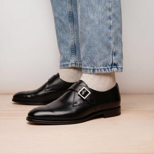 Può includere: Un paio di scarpe in pelle nera con cinturino a fibbia singola, fibbia in metallo argentato, punta arrotondata e perforazioni decorative brogue sulla punta. Le scarpe sono indossate con calzini a coste chiari e jeans azzurri.