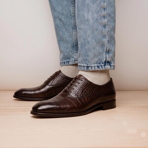 May include: A pair of dark brown leather oxford shoes featuring a textured crocodile skin pattern, thin brown laces, and a low heel. The shoes are worn with light beige ribbed socks and light blue denim jeans, showcasing a classic lace-up design.