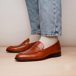 Op de afbeelding: Een paar cognacbruine leren penny loafers met een klassieke band over de wreef en een lage hak. De schoenen worden gedragen met lichtbeige geribbelde sokken en een lichtblauwe spijkerbroek, met een gladde leren afwerking en stiksels.