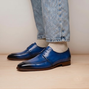 May include: A pair of blue leather derby shoes with a polished finish, featuring a dark gradient at the toe, thin black laces, and a brown stacked heel. The shoes are worn with light-wash denim jeans and beige socks, showcasing a classic silhouette.