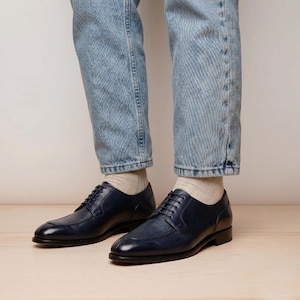 Peut inclure: Une paire de chaussures de ville en cuir bleu marine foncé avec un bout texturé, des lacets noirs fins et un petit talon, portées avec des chaussettes côtelées beige clair et un jean bleu clair s'arrêtant juste au-dessus de la cheville.