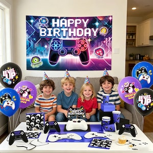 Op de afbeelding: Een verjaardagsfeestset met een videogame-thema, bestaande uit een grote achtergrond met een neoncontroller, ballonnen met game-afbeeldingen, feesthoedjes, een taarttopper, papieren bekers en controller-vormige decoraties op een witte tafel.