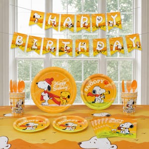Op de afbeelding: Een Peanuts thema feestset met oranje papieren borden, bekers en servetten versierd met illustraties van Snoopy en Woodstock. De set bevat een Happy Birthday slinger met witte letters en bijpassende oranje plastic vorken en lepels.
