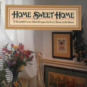 May include: A book cover titled "Home Sweet Home" featuring a framed title and the subtitle "25 Beautiful Cross-Stitch Designs for Every Room in the House." The cover displays a floral arrangement, framed artwork, and a table setting.