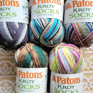 May include: A collection of six skeins of Patons Kroy Socks yarn in various color combinations. The yarn labels are white with green and orange text. The yarn colors include shades of blue, gray, brown, green, and pink. Each skein is 50 grams.