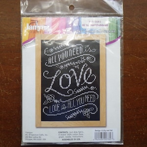 May include: A cross-stitch kit featuring a black chalkboard with white lettering that reads "All you need is love, Love is all you need". The kit includes Aida fabric, floss, a needle, and instructions. It is assembled in the USA.