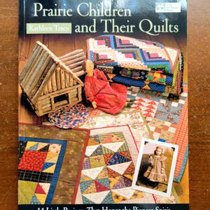May include: A softcover book titled Prairie Children and Their Quilts by Kathleen Tracy. The cover features a small log cabin model, a doll in a red dress, several colorful patchwork quilts, and a vintage photograph of a young child.