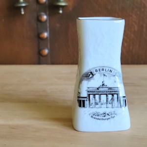May include: White ceramic vase with a black and white illustration of the Brandenburg Gate in Berlin. The vase has a rectangular shape with a slightly curved design and the word "BERLIN" at the top of the illustration.