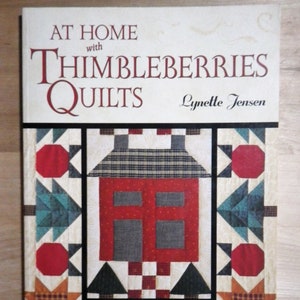 May include: A book cover titled "At Home with Thimbleberries Quilts" by Lynette Jensen. The cover features a quilt design with a red house, green trees, and star patterns. The book is a collection of 25 country quilts and decorative accessories.