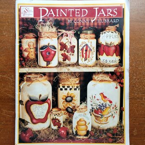 May include: A craft book titled Painted Jars by Conny Hubbard featuring a collection of white glass jars decorated with hand-painted designs including apples, sunflowers, a beehive, a birdhouse, and a gingerbread person, all arranged on shelves.