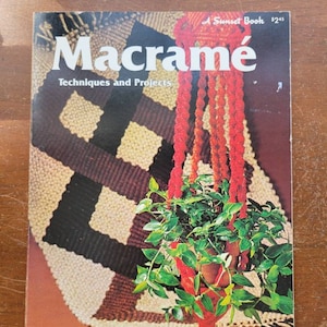 May include: A vintage book titled "Macramé Techniques and Projects" with a revised edition. The cover features a macramé plant hanger holding a potted plant with green leaves. The background shows a macramé wall hanging with brown, black, and beige patterns.