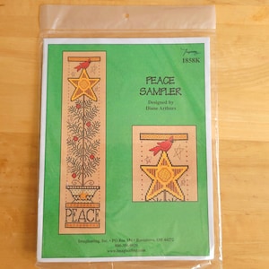 May include: A cross-stitch sampler kit in a plastic sleeve. The design features a star, a bird, and the word "PEACE". The kit is designed by Diane Arthurs. The background is green with a tan-colored design.