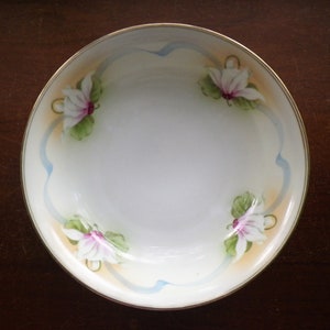 May include: A white porcelain bowl with a gold rim, featuring hand-painted floral designs. The flowers are white with pink centers and green leaves, accented by a light blue ribbon and a yellow-orange background.